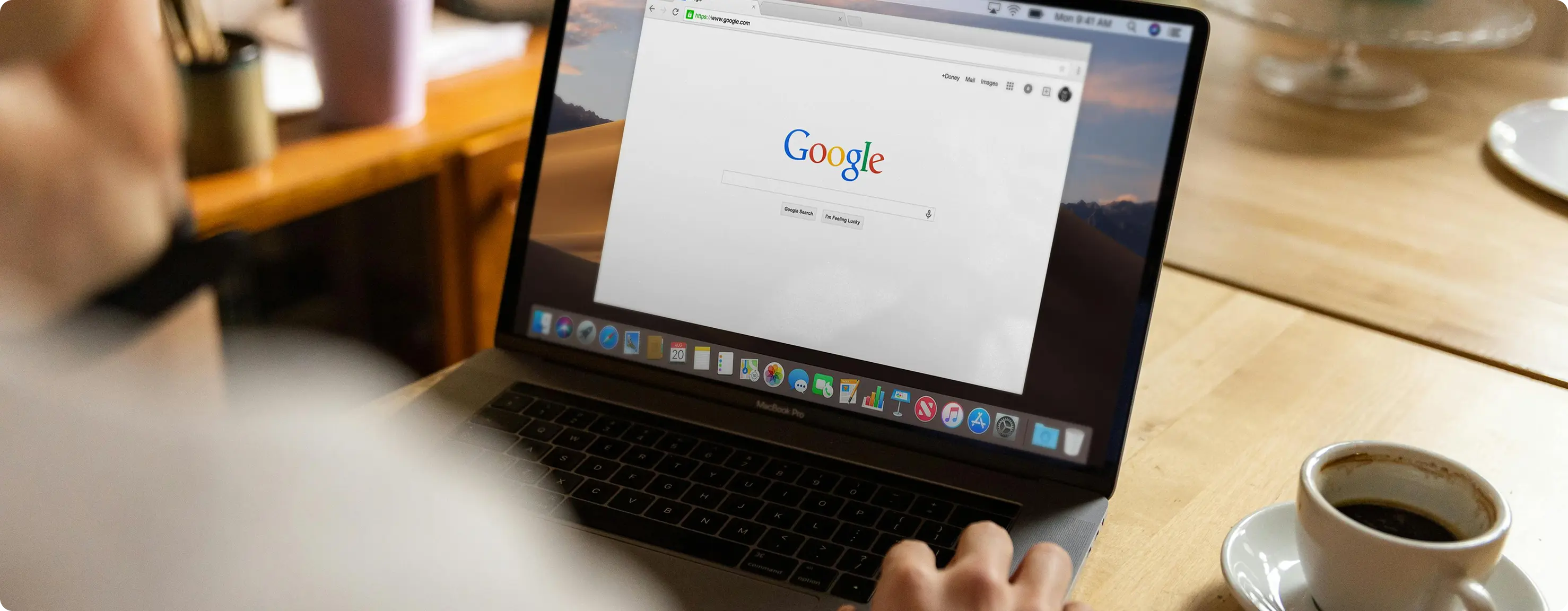 A person using a MacBook Pro at a wooden table with a cup of coffee, viewing the Google search homepage on the screen.